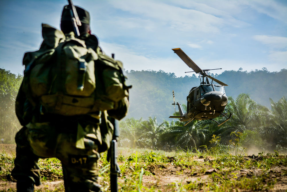 A helicopter lands in a jungle clearing as a soldier looks on.